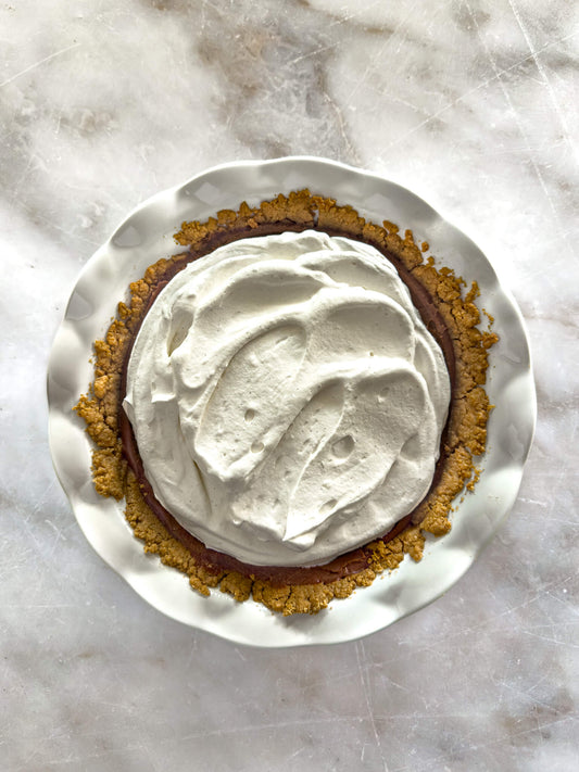 Gluten-free chocolate silk cream pie in a white ruffled-edge pie plate, topped with Madagascan vanilla whipped cream.